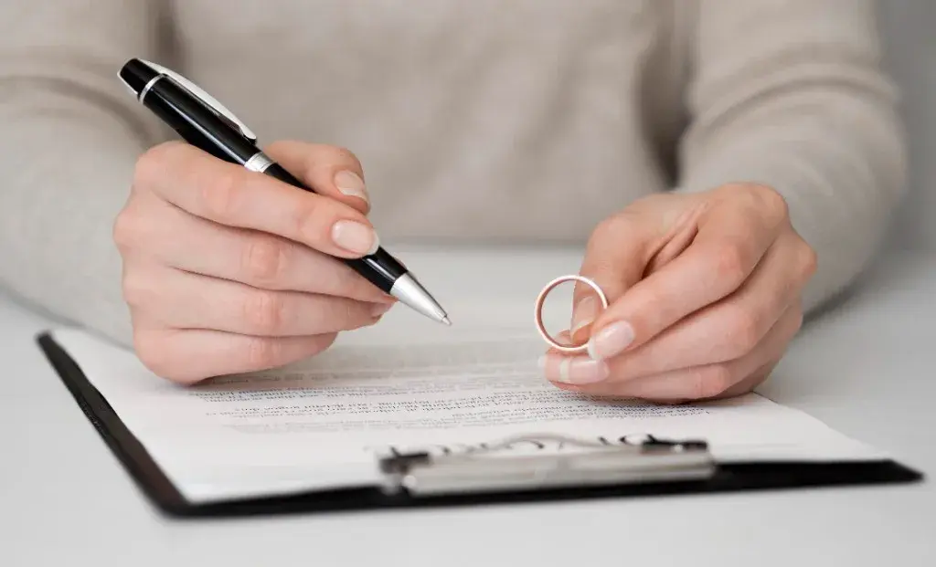 Hands holding a wedding ring and pen over a divorce document on a clipboard